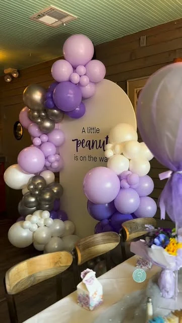 Reel-cover still of lavender, ivory and gold balloons around a rounded baby shower backdrop.