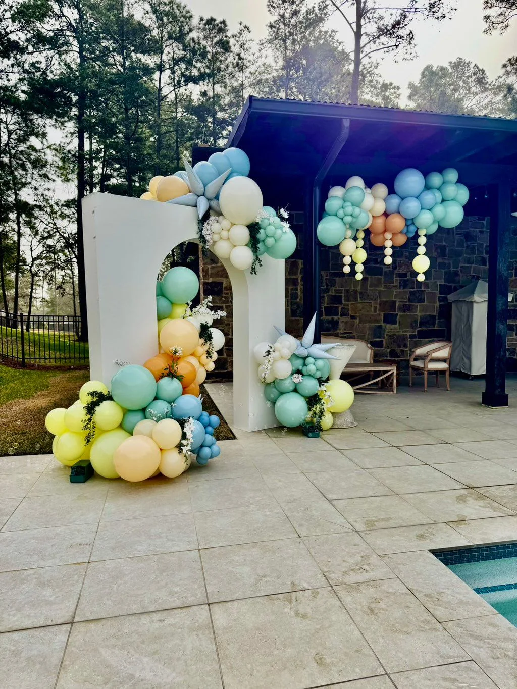 Pastel yellow, blue, mint and white balloons surround outdoor panels, with hanging accents beneath a covered patio.
