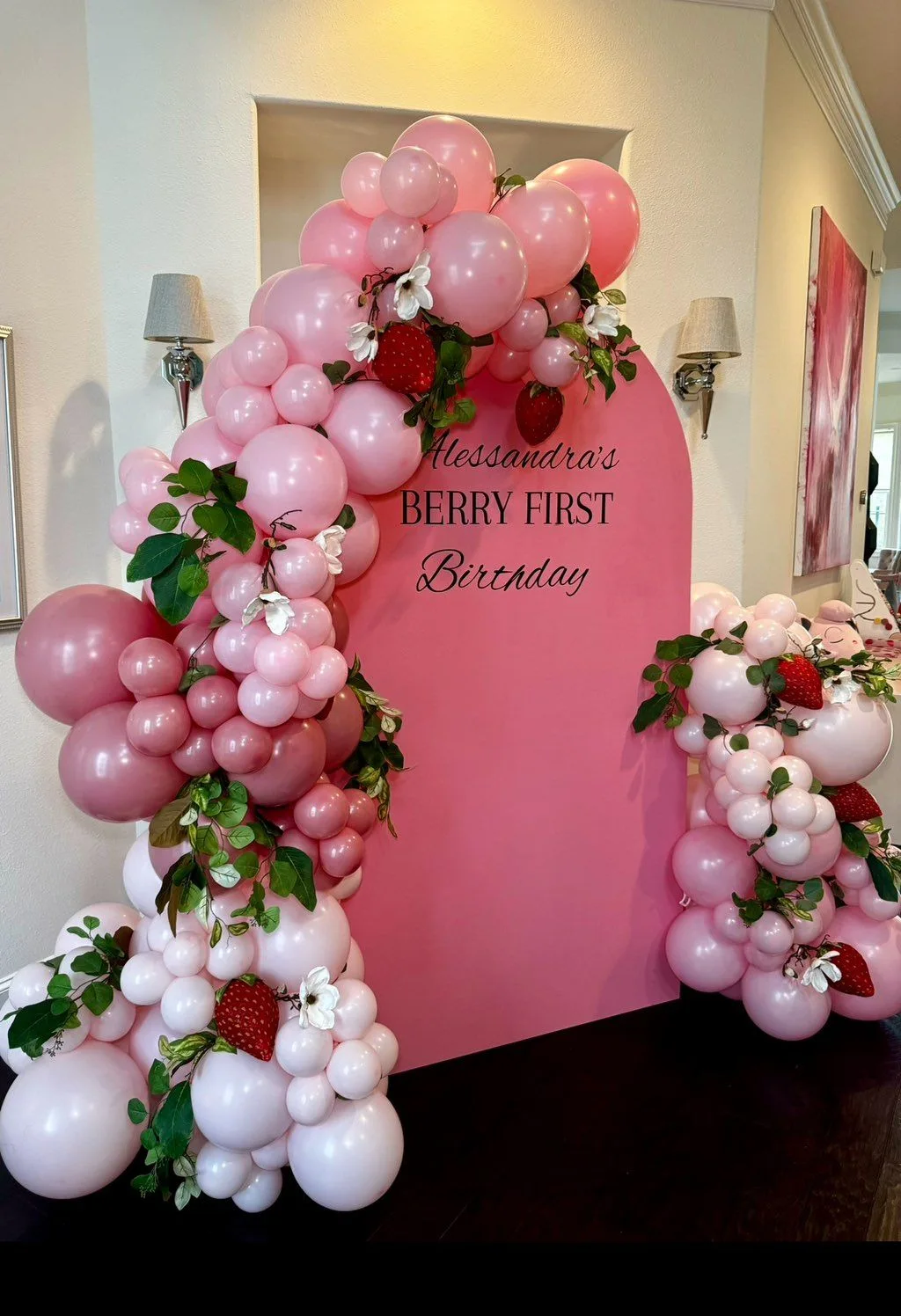 Pink, white and red balloons with strawberry and floral details surround a first-birthday backdrop.